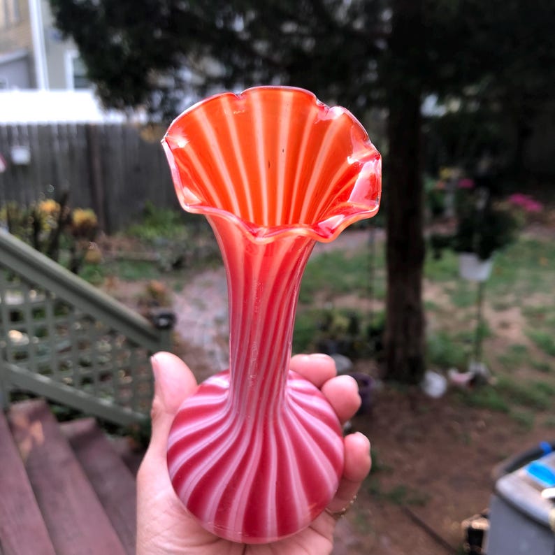 May include: A hand holds a vintage glass vase. The vase has a ruffled rim and a long, slender neck. The vase is red and white with a striped pattern. The base of the vase is round.