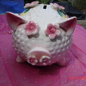 May include: White ceramic piggy bank with pink flowers and a white polka dot pattern. The piggy bank has a slot on the top for coins.