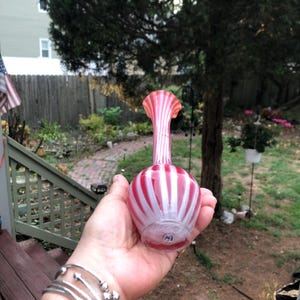 May include: A hand holds a decorative glass vase. The vase has a bulbous base and a tall, fluted neck. It features alternating vertical stripes of red and white. The vase is likely vintage and adds a touch of elegance.
