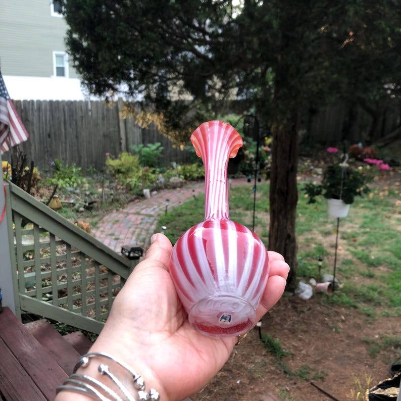 May include: A hand holds a decorative glass vase. The vase has a bulbous base and a tall, fluted neck. It features alternating vertical stripes of red and white. The vase is likely vintage and adds a touch of elegance.