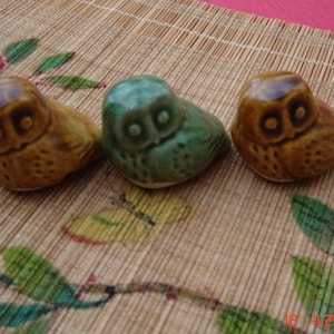 May include: Three ceramic owl figurines in shades of brown and green are arranged on a bamboo mat with floral accents. The owls have large, round eyes and are in a sitting position. The figurines are approximately 2 inches in height.