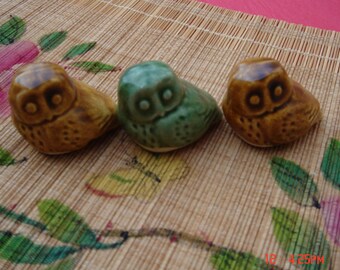 May include: Three ceramic owl figurines in shades of brown and green are arranged on a bamboo mat with floral accents. The owls have large, round eyes and are in a sitting position. The figurines are approximately 2 inches in height.