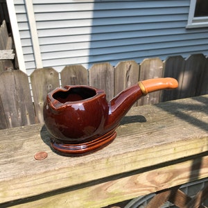 May include: A brown ceramic pipe-shaped ashtray with a brown glazed finish and a light brown handle.