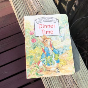 May include: A children's book titled "Dinner Time" by Beatrix Potter. The cover features an illustration of a rabbit in a blue jacket holding carrots. The book is set against a backdrop of a garden with flowers and greenery.