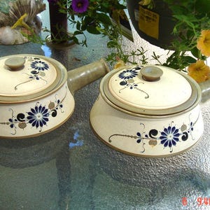 May include: Two ceramic casserole dishes with lids. The dishes are white with a brown rim and blue and gold floral designs. The lids have a knob on top.