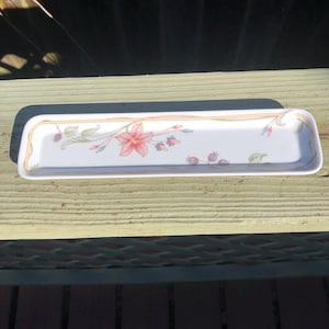 May include: A white ceramic serving dish with a floral design. The dish has a gold trim and features a pink flower with green leaves and purple berries.
