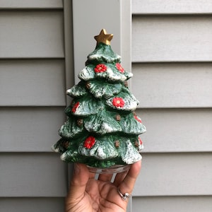 May include: A small, ceramic Christmas tree figurine with a green and white frosted finish. The tree is decorated with red flowers and a gold star on top.