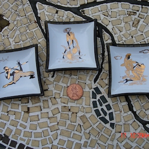 May include: Three small, square, white ceramic dishes with black trim. Each dish features a gold design with a different scene. The first dish has a person bending backwards in a limbo pose. The second dish has a person holding a fish. The third dish has a person playing a musical instrument. The text "Limbo!" is on the first dish.
