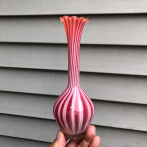 May include: A tall, slender vase with a bulbous base and a ruffled rim. The vase is striped with alternating bands of red and white. The vase is held in a person's hand.