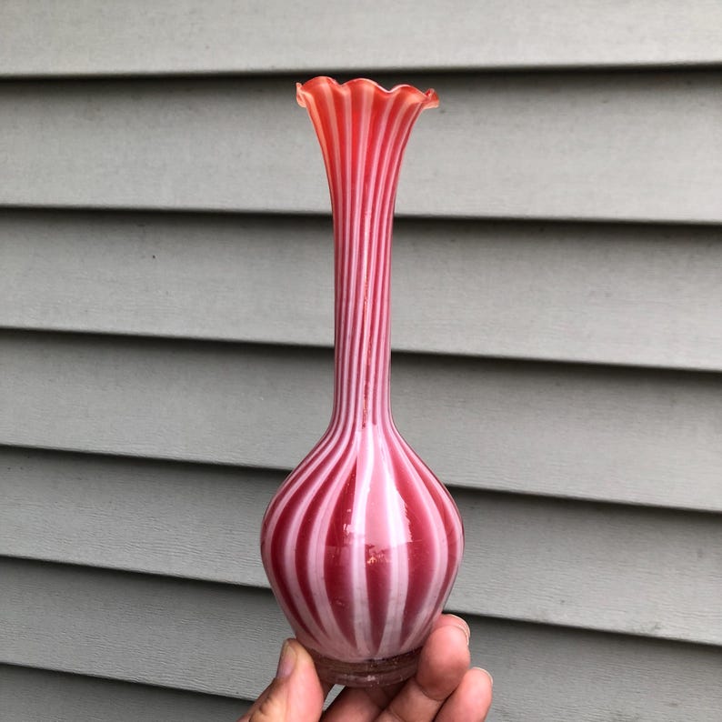 May include: A tall, slender vase with a bulbous base and a ruffled rim. The vase is striped with alternating bands of red and white. The vase is held in a person's hand.