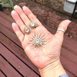 May include: A set of vintage sunburst earrings and brooch with clear rhinestones. The jewelry is gold-toned with silver accents.