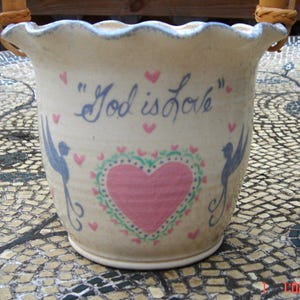 May include: A white ceramic pot with a scalloped rim and two brown leather handles. The pot is decorated with blue birds, pink hearts, and green leaves. The text "God is love" is written in blue on the front of the pot.