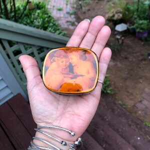 May include: A rectangular brooch with rounded corners featuring a warm-toned, amber-colored lacquer finish. The surface displays a detailed illustration of two winged cherubs with golden hair, positioned closely together while looking downward.