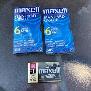 May include: Two blue and white Maxell VHS video cassette tapes labeled "Standard Grade" with a 6 hour recording time in EP mode.  A Maxell XLII audio cassette tape with a black and white image of a person in a car.