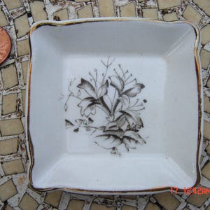 May include: A white porcelain dish with a gold rim and a black floral design. The dish is square and has a raised edge.