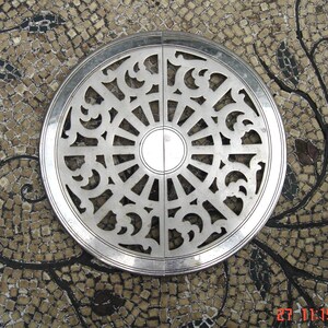 May include: A silver, circular decorative object with an openwork design featuring symmetrical patterns. The object is set against a mosaic background with a natural, earthy color palette. The design includes a central circle and radiating lines.