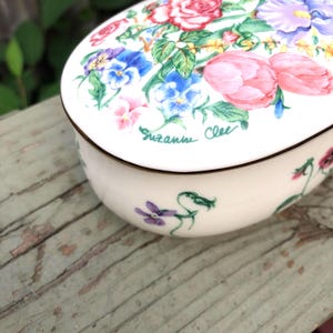 May include: A white ceramic trinket box with a floral design. The lid features a bouquet of pink, purple, and blue flowers with green leaves. The box is signed "Suzanne Clea".