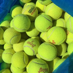 May include: A blue plastic bag filled with yellow tennis balls. The balls are various brands, including Wilson US Open and Penn.
