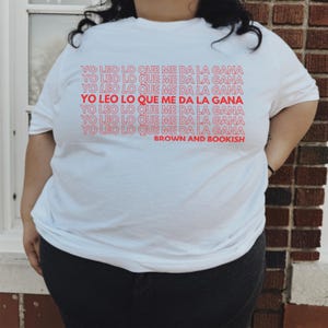 Könnte beinhalten: Weißes T-Shirt mit dem Satz "Yo leo lo que me da la gana", der mehrmals in roter Schrift wiederholt wird. Der Satz ist in einem Retro-Schriftart-Stil. Der Text "Brown and Bookish" ist unter dem wiederholten Satz gedruckt.