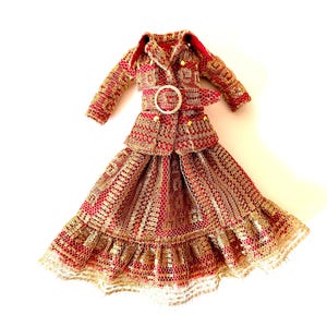 May include: A miniature red, gold, and white patterned skirt suit with a matching jacket and skirt. The jacket has a belt with a white circular buckle. The skirt has a ruffled hem.