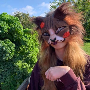 Pode incluir: Uma máscara de gato castanha e branca com pelo sintético longo e texturizado, orelhas pontiagudas e marcas faciais estilizadas. A máscara inclui detalhes em laranja perto dos olhos e orelhas, um nariz escuro e uma boca visível.