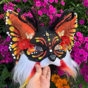 Puede incluir: Una máscara de zorro con patrones de alas de mariposa naranjas y negras en las orejas, cara negra con marcas amarillas y naranjas, mejillas de piel sintética blanca, flores de tela rojas, pequeños detalles de mariposas doradas y nariz negra.