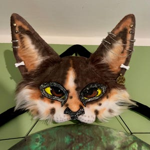 Può includere: Una maschera animale in pelliccia sintetica con macchie marroni, bianche e arancioni e grandi occhi gialli e marroni. Le orecchie sono decorate con piercing ad anello argentati, stelline, ciondoli a forma di osso, una catenina e un ciondolo a zampa.