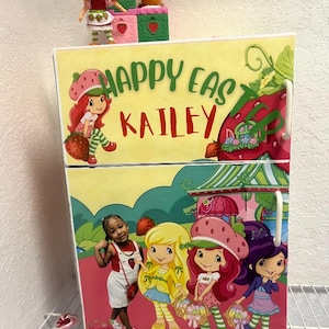 May include: A pink and green toy refrigerator with a Strawberry Shortcake theme. The refrigerator has a handle on each door and a sticker that says "Happy Easter Kailey". The refrigerator is decorated with images of Strawberry Shortcake and her friends.