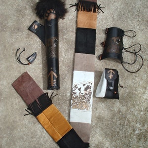 K&ouml;nnte beinhalten: Ein brauner Lederk&ouml;cher mit einem B&auml;renpfoten-Design, ein brauner Lederarmschutz und ein brauner Lederpfeilhalter mit einem B&auml;ren-Design. Der K&ouml;cher ist mit Fransen verziert und hat einen wei&szlig;en und braunen Stoffgurt mit einem B&auml;ren-Design.