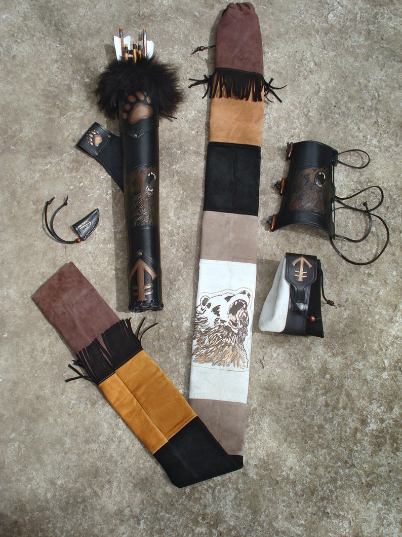 K&ouml;nnte beinhalten: Ein brauner Lederk&ouml;cher mit einem B&auml;renpfoten-Design, ein brauner Lederarmschutz und ein brauner Lederpfeilhalter mit einem B&auml;ren-Design. Der K&ouml;cher ist mit Fransen verziert und hat einen wei&szlig;en und braunen Stoffgurt mit einem B&auml;ren-Design.