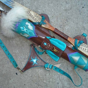 May include: A handcrafted archery set featuring a brown leather quiver with teal and purple accents, a matching arm guard, and a bow. The quiver has a white fur trim and holds arrows. The set is designed with intricate details.