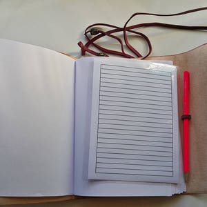 Może przedstawiać: Otwarty brązowy notes sk&oacute;rzany z r&oacute;żowym długopisem i liniowanym papierem w środku. Notes ma sk&oacute;rzany pasek zamykający.
