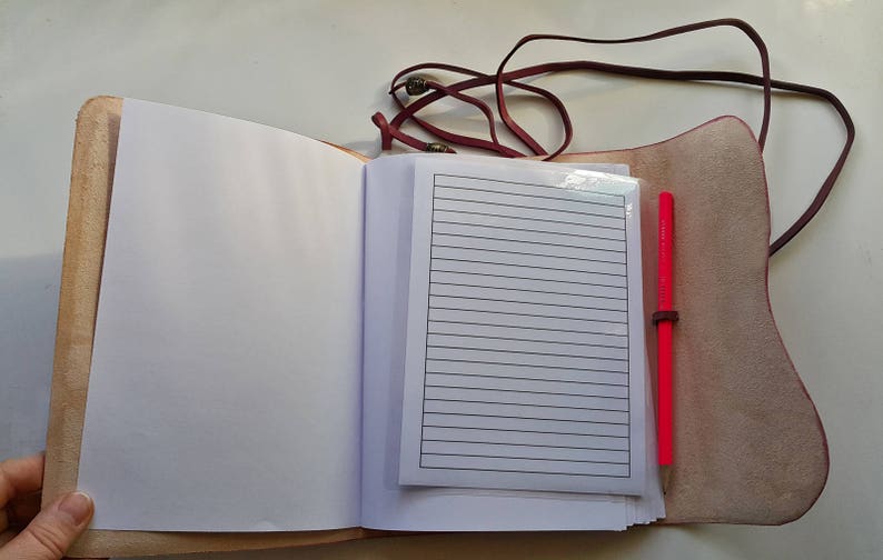 Może przedstawiać: Otwarty brązowy notes sk&oacute;rzany z r&oacute;żowym długopisem i liniowanym papierem w środku. Notes ma sk&oacute;rzany pasek zamykający.
