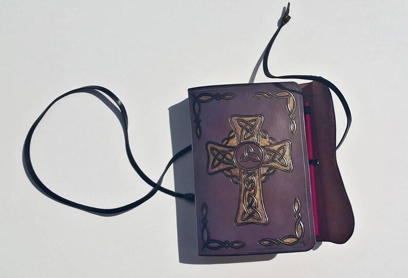 Op de afbeelding: Een paars lederen dagboek met een gouden Keltisch kruisontwerp en een bijpassende penhouder. Het dagboek heeft een zwarte band sluiting en een bruine lederen flap.