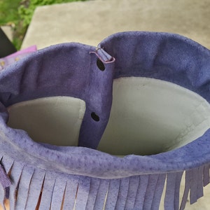 K&ouml;nnte beinhalten: Lilafarbene Wildledertasche mit Fransenbesatz und Druckknopfverschluss. Die Tasche ist mit hellem grauen Stoff gef&uuml;ttert.