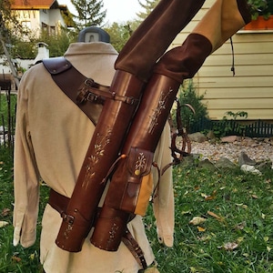 Op de afbeelding: Een bruine leren pijlkoker met twee vakken voor pijlen, gedragen op de rug van een persoon met een schouderriem en een riem. De pijlkoker heeft een decoratief bloemenpatroon en een klein zakje aan de zijkant.