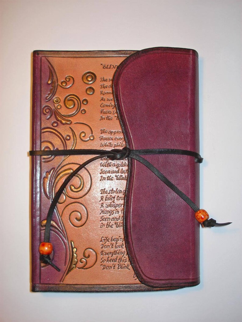 Op de afbeelding: Een bordeauxrood lederen dagboek met een tan lederen omslag met een decoratief bloemmotief en een gedicht met de titel "BLIND". Het dagboek is gesloten en vastgebonden met een zwart lint.