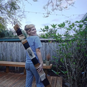 May include: A woman wearing blue pants and a blue shirt is holding a long, brown and black carrying case with a white tassel. The case has a black and white design on it.