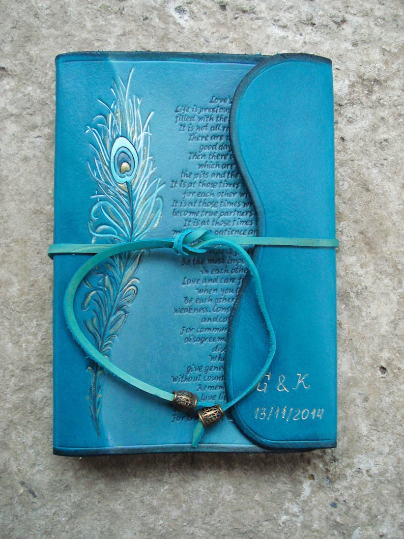 Op de afbeelding: Een tealblauw lederen dagboek met een gegraveerd pauwenveerontwerp op de voorzijde. Het dagboek heeft een citaat over liefde en geduld dat op de voorzijde is gegraveerd. Het dagboek is vastgebonden met een tealblauwe lederen band en heeft de initialen "G&K" en de datum "13/11/2014" gegraveerd op de voorzijde.