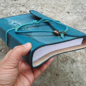 Op de afbeelding: Turkoois lederen gebonden dagboek met een pauwenveerontwerp en een leren riemsluiting. Het dagboek heeft blanco pagina's en is gesloten.