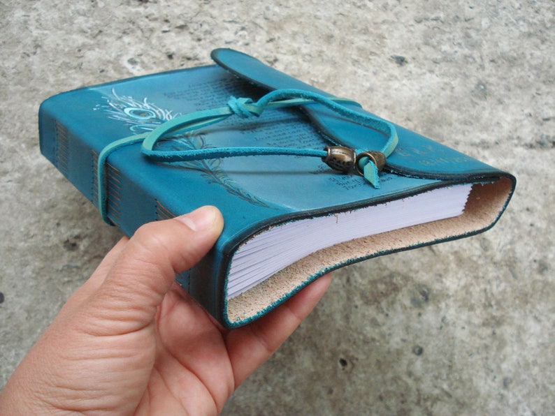 Op de afbeelding: Turkoois lederen gebonden dagboek met een pauwenveerontwerp en een leren riemsluiting. Het dagboek heeft blanco pagina's en is gesloten.