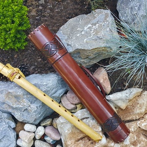 May include: A wooden flute with a brown leather carrying case. The case features a Celtic knot design. The flute is resting on a bed of rocks.