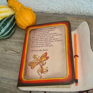 Może przedstawiać: Sk&oacute;rzany notes z cytatem Winniego the Pooha o odwadze, sile i inteligencji. Notes ma wz&oacute;r ważki i jasnopomarańczowy oł&oacute;wek włożony w oprawę.