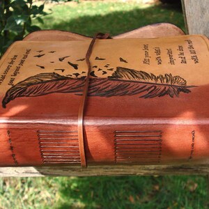 Op de afbeelding: Een bruin lederen dagboek met een verenontwerp en de quote "What is cover for love goes beyond good and evil."  Fit your journey with what's important. And be the wind while all the rest...