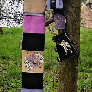 K&ouml;nnte beinhalten: Ein violetter, schwarzer und brauner Lederk&ouml;cher mit wei&szlig;em Pelzbesatz und einem kleinen violetten Lederbeutel. Der K&ouml;cher h&auml;ngt an einem Baumzweig.