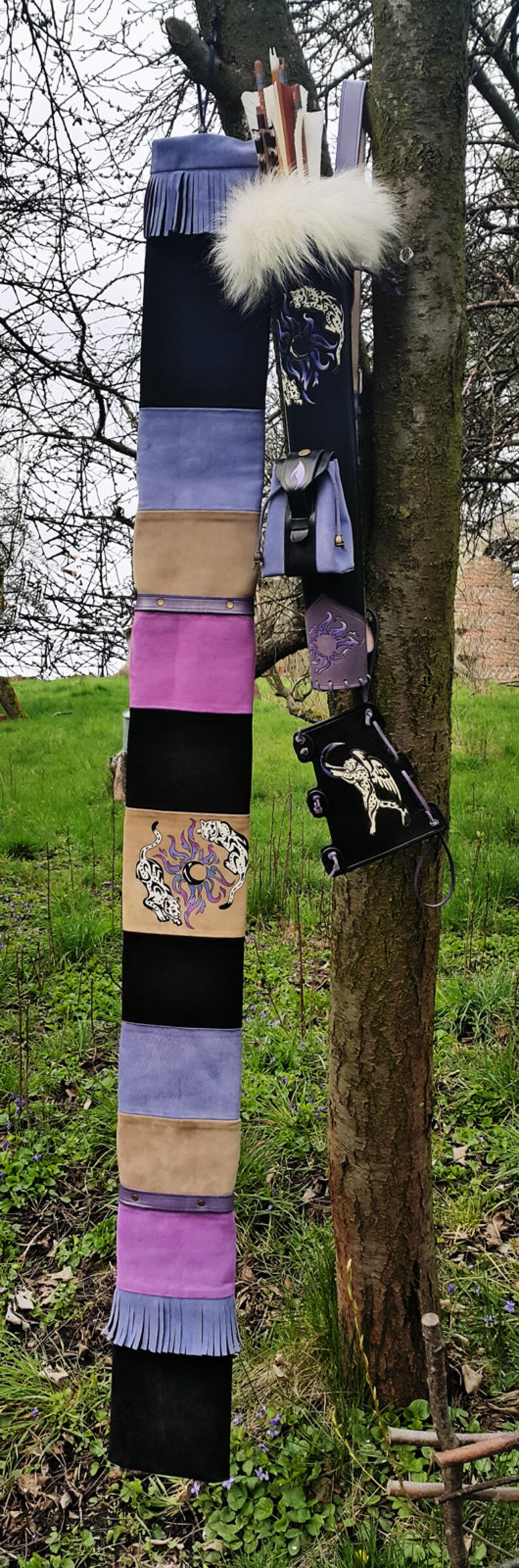 K&ouml;nnte beinhalten: Ein violetter, schwarzer und brauner Lederk&ouml;cher mit wei&szlig;em Pelzbesatz und einem kleinen violetten Lederbeutel. Der K&ouml;cher h&auml;ngt an einem Baumzweig.