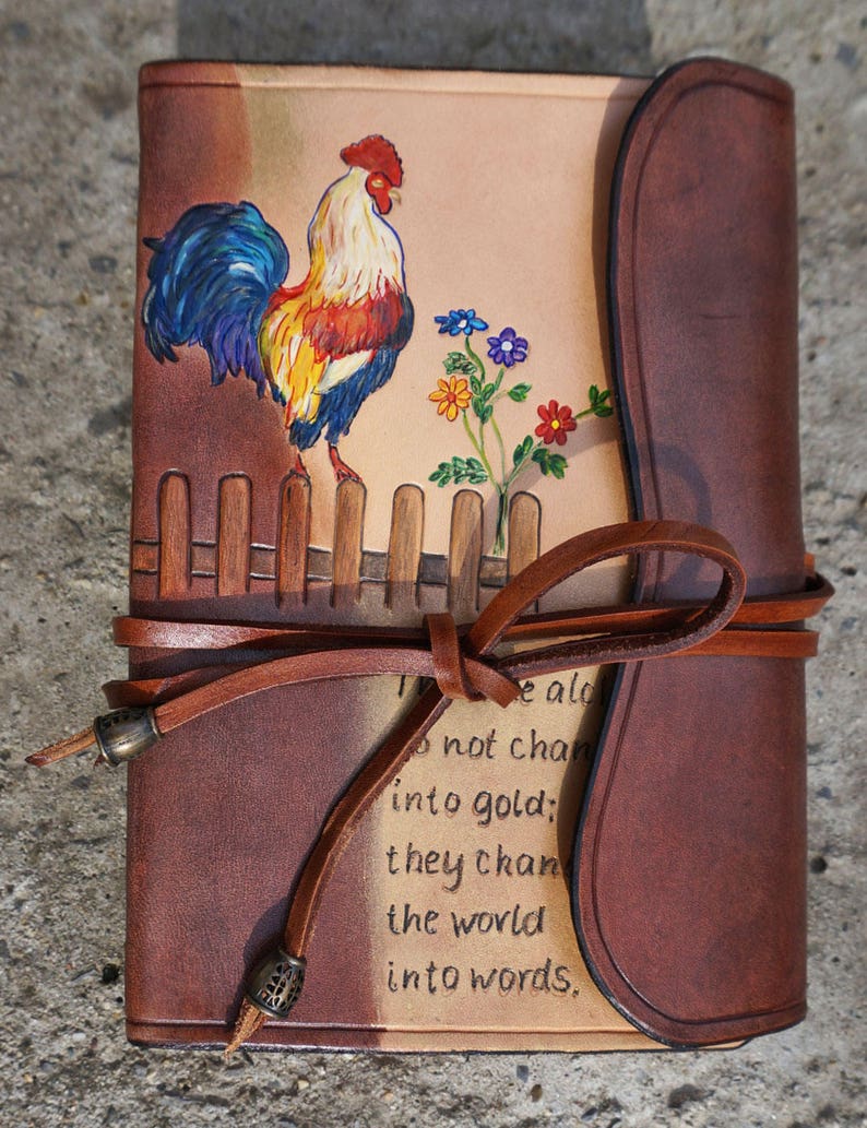 Może przedstawiać: Brązowy sk&oacute;rzany dziennik z namalowanym na okładce kogutkiem i kwiatami. Dziennik ma sk&oacute;rzany pasek z metalowymi akcentami i cytat, kt&oacute;ry brzmi: "Nie zmieniają się w złoto; zmieniają świat w słowa."