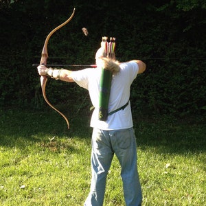 May include: A person wearing a white shirt and blue jeans is holding a bow and arrow. The bow is made of wood and has a string attached to it. The arrow is made of wood and has a metal tip. The person is standing in a grassy field.