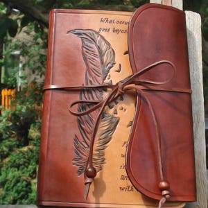 Op de afbeelding: Een bruin leren dagboek met een gegraveerd ontwerp van een veer en de tekst "What occurs goes beyond..." Het dagboek heeft een leren riemgesp en een flap die de voorkant bedekt.