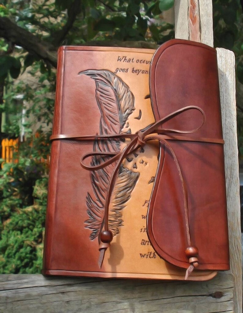 Op de afbeelding: Een bruin leren dagboek met een gegraveerd ontwerp van een veer en de tekst "What occurs goes beyond..." Het dagboek heeft een leren riemgesp en een flap die de voorkant bedekt.